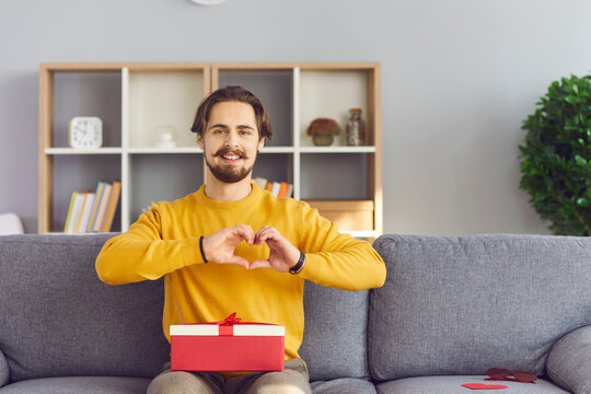 Smiling young man forming heart fron fingers and holding holiday present on knees during online dating on sofa at home. Valentines day, holiday gift, anniversary, dating, congratulation, date concept