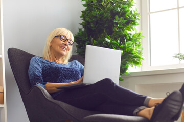Mature woman with glasses sitting in an easy chair and working at home using a laptop. Senior woman doing business remotely sitting in a comfortable home office. Online business concept.