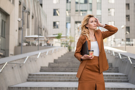 Beautiful blonde woman in brown classic smart-casual outfit outdoors near hi-tech business building. She drinks coffee in to go cup and enjoy summer warmth. Space for text.