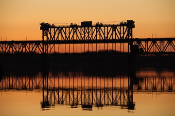 Fototapeta premium Traffic on the bridge over the Dnieper river against the background of an orange sunset. Kryukovsky road-rail bridge in Kremenchug, Ukraine. Industrial silhouette against the background of a natural