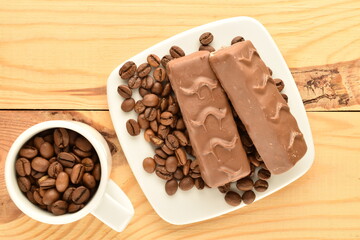 Two chocolate energy bars, a cup and a saucer with coffee grains, close-up, on a wooden table.