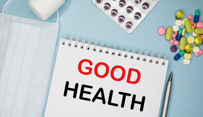 GOOD HEALTH is written in a notebook on a blue background next to pills, mask and pen.