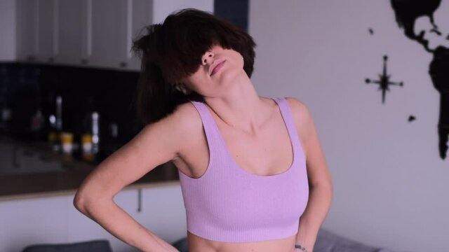 Sleepy woman at home in the morning in an apartment in a sporty lilac top and leggings does exercises, kneads joints, neck, shoulders. Tattoo on back in thai language means "Enjoy your life"  
