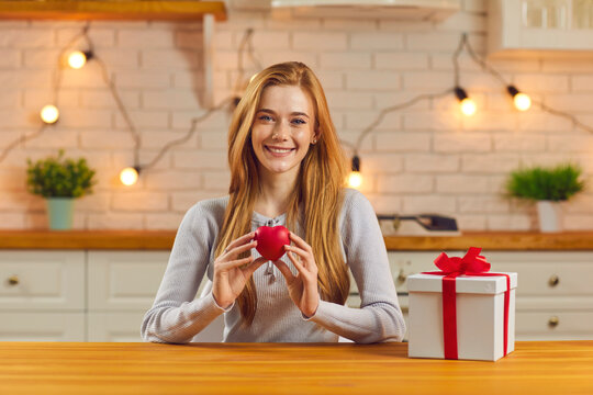 Remote love. Redhead woman sitting at home in the kitchen at the table near the gift and shows a red heart to the webcam. Young girl is talking on a video call with her lover on Valentine's Day.