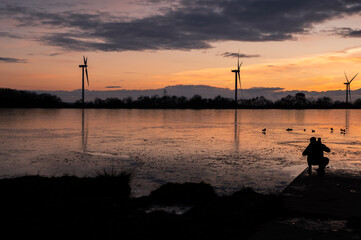 Shadow of young male overlooking frozen lake with child with turbines in the background duing sunset. south wales uk.