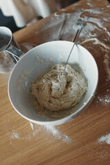 White bowl with dough and spoon in it, on the wooden table in the kitchen. The process of mixing ingredients for pastry. Closeup, selective focus.