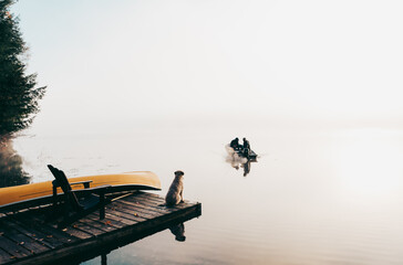 Two people in fishing boat in the fog with dog watching from the dock.