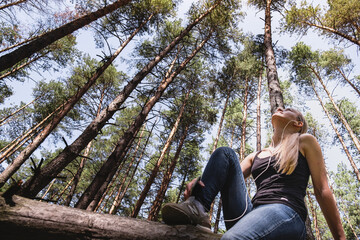 a girl in a forest sits on a tree, with headphones listening to music. rest at nature. in the background blurred silhouettes of trees. digital detox. Mental health. Positive emotion.