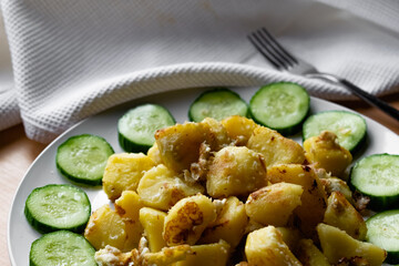 Fried potato slices in a beaten egg with protein, around a circle of green juicy fresh cucumber. Dish on a white plate on a wooden table