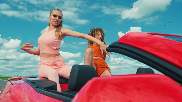 Two young beautiful hipster girls in red convertible car. Sexy carefree women in a cabriolet. Positive models is riding and having fun. Raising hands at summer and blue sky on background.