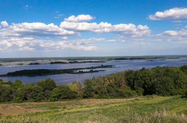 Below, under the hill, there is a wide river against the background of light clouds and a bright blue sky