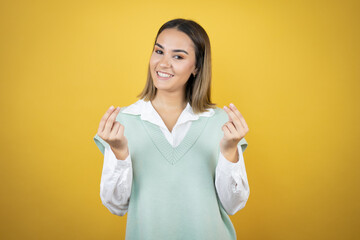 Pretty young woman standing over yellow background doing money gesture with hands, asking for salary payment, millionaire business