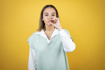 Pretty young woman standing over yellow background with mouth and lips shut as zip with fingers....