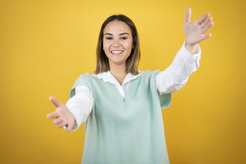 Pretty young woman standing over yellow background looking at the camera smiling with open arms for hug