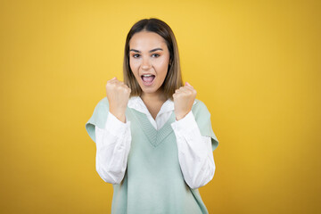 Pretty young woman standing over yellow background very happy and excited making winner gesture with raised arms, smiling and screaming for success.