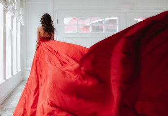 Brunette woman with long hair standing by her back in long red evening dress in white room.