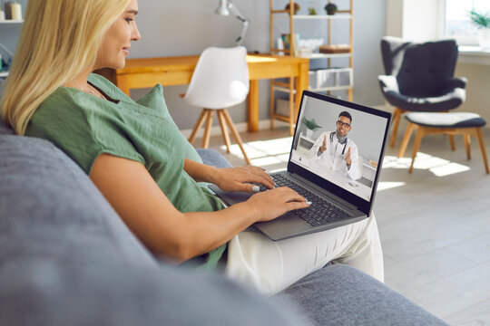 Telemedicine, E-health Clinic, Virtual Visit To Hospital In Times Of Pandemic Concept. Happy Young Woman Sitting On Sofa In Living-room And Talking To Online Doctor Via Video Call On Laptop Computer