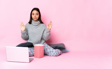 Young mixed race woman eating popcorn while watching a movie on the laptop making stop gesture and disappointed