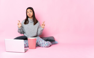 Young mixed race woman eating popcorn while watching a movie on the laptop with fingers crossing and wishing the best