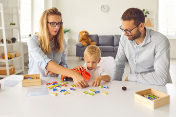 Focused mom, dad and toddler son play and compose words from scattered colored letters. Family sits...