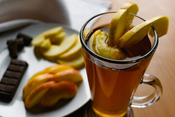 Apple tea in a transparent glass with a handle, the drink is decorated with slices of fresh apple with a yellow peel, next to the plate of sliced fruit and chocolate, everything is on the table on a w