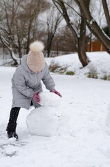 Fototapeta na wymiar A girl in winter clothes and a hat playing with snow in a winter park. The child is outside during the winter holidays. A child makes a snowman with snow.
