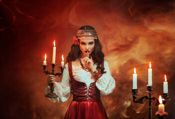 Fantasy gypsy woman in red vintage dress. Long black hair, gold jewelry. Girl witch fortune teller holds a candlestick with burning candles. Room is on fire, smoke. Spiritual seance, summons spirits. - obrazy, fototapety, plakaty