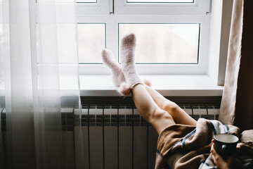 Young woman resting on armchair near the window, drinking hot tea wrapped in a blanket.