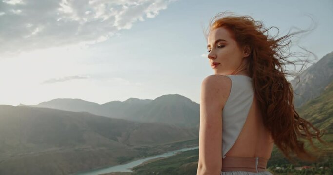 Beautiful caucasian girl with red hair posing for a photoshoot on top of a mountain. Young model on scenic background - freedom, real people 4k footage