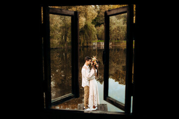 Loving couple in festive attire standing in an embrace on the pier on the background of the lake, view from the window of the house