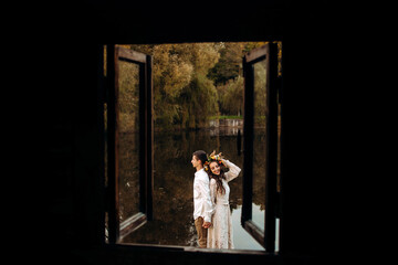 Loving couple in festive attire standing in an embrace on the pier on the background of the lake, view from the window of the house