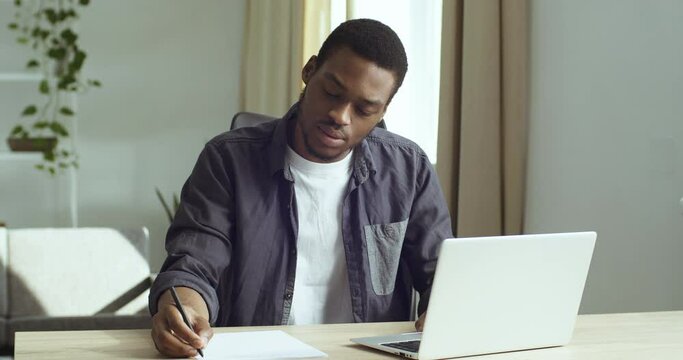 Young guy student pensive afro american business man freelancer sits at table in front of laptop works studies writes with pen on white paper in document plans analyzes does paper work in home office 