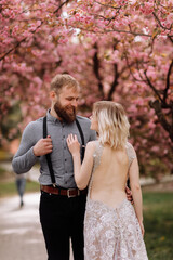 Fototapeta premium Beautiful, cheerful and lively newlyweds, groom and bride are hugging near the blooming pink cherry blossom. Wedding portrait of a close-up of a smiling bearded groom and a cute bride.