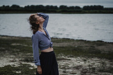 Moody vibe portrait of a young caucasian woman in large pants and blue striped shirt, holding hair looking up,  on a rural lake background 