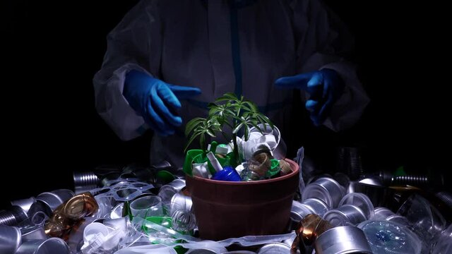 Environmentalist in protective suit holding young seedling in pot full of plastic dirt trash. Concept and symbol of catastrophic apocalypse, eco sustainability, growth care, protecting the earth.