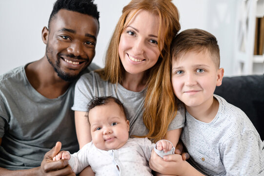 A portrait of happy multiracial family at home. A multiethnic spouses, their cute newborn baby girl and school-age son look into the camera and smile.