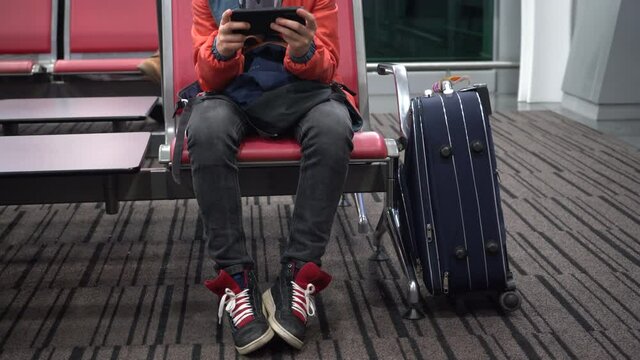 A teenage passenger with luggage is waiting for his flight at the airport and uses a smartphone.