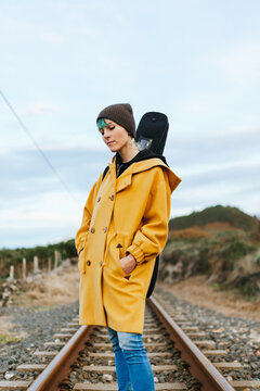 Young woman with yellow coat and serene gesture looking at the ground on the train tracks with the guitar loaded on her back and with her hands in her pockets. wanderluster. youth and free time