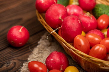 Green cucumbers, ripe radishes and colorful tomatoes in a wicker basket and scattered on the table. Close-up, selective focus.