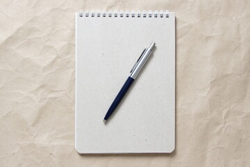 Gray notepad with white coiled spring and pen on a background of beige crumpled craft paper. With empty space for text and design