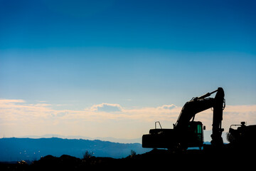 Backlit silhouette of an excavator on top of a hill with unfocused sky background.