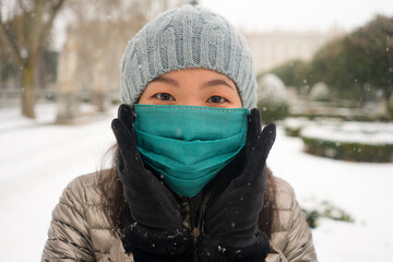 Fototapeta na wymiar snowfall in covid19 time - young happy and attractive Asian Japanese woman in winter hat and mask at beautiful city park playing with snow cheerful enjoying cold Christmas holidays