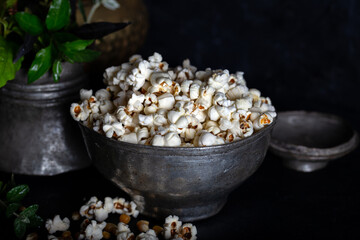 Prepared popcorn in frying pan, corn seeds in bowl and corncobs on dark background.