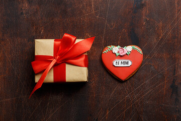 Cardboard gift box tied with a red satin ribbon and red heart shaped gingerbread cookie on vintage wooden background. Overhead view, copy space. Valentine's Day concept