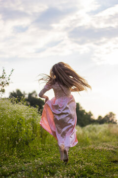 Back View Of Woman With Long Blonde Hair Running Across The Wildflowers Field Barefoot Holding The Bottom Of The Long Pink Dress On A Warm Sunny Day. Joyfull Summer Activities. Girl From Wonderland.