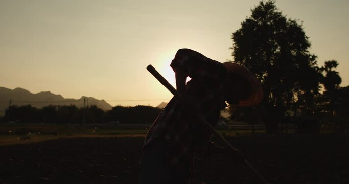 Slow motion silhouette scene of a rural Asian farmer stands with a hoe over his shoulder as the sun sets. With sunlight passing through him beautifully.