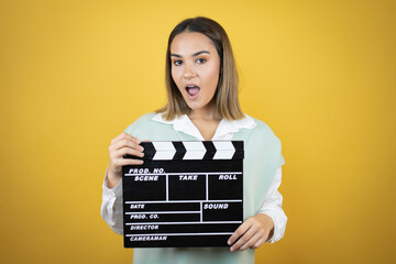Pretty young woman standing over yellow background holding a clapperboard
