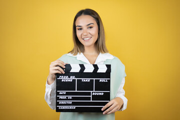 Pretty young woman standing over yellow background holding a clapperboard