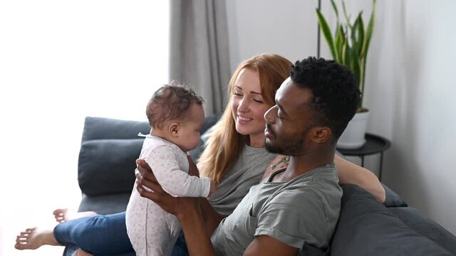 An African-American father throws his cute baby girl up, a beautiful caucasian mother sits on the couch near. Young multiracial parents enjoy time together with newborn child at cozy home, they smile