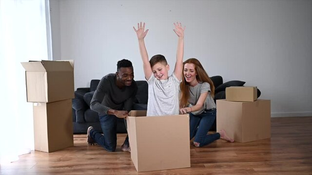 A multiracial family have fun in a new house, an African stepfather and caucasian mother driving school-age son in a cardboard box, they are laughing. Concept of moving in a new house